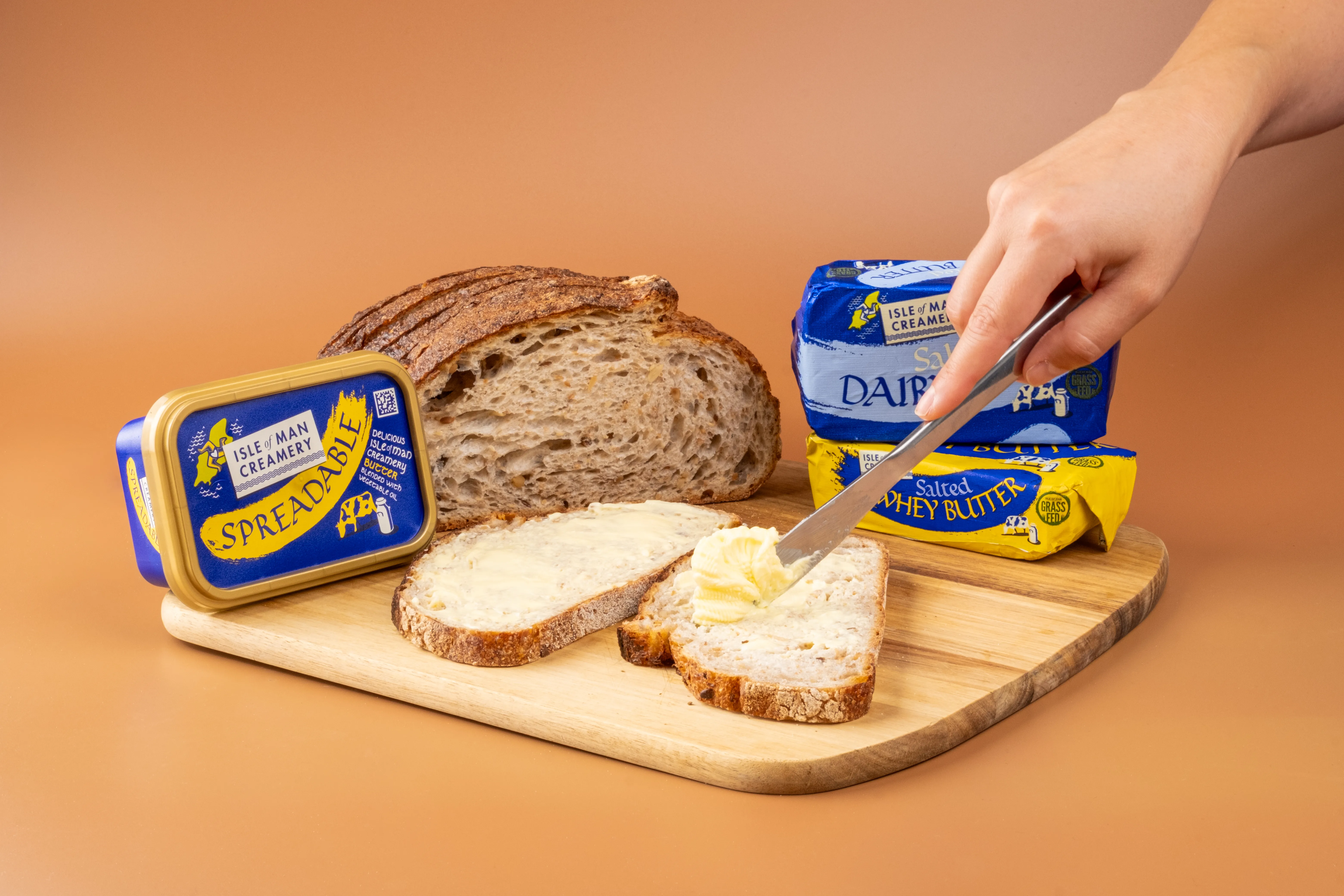 A hand spreading Isle of Man Creamery spreadable butter onto sliced sourdough bread on a wooden chopping board, with a butter pat in the background, on an orange background